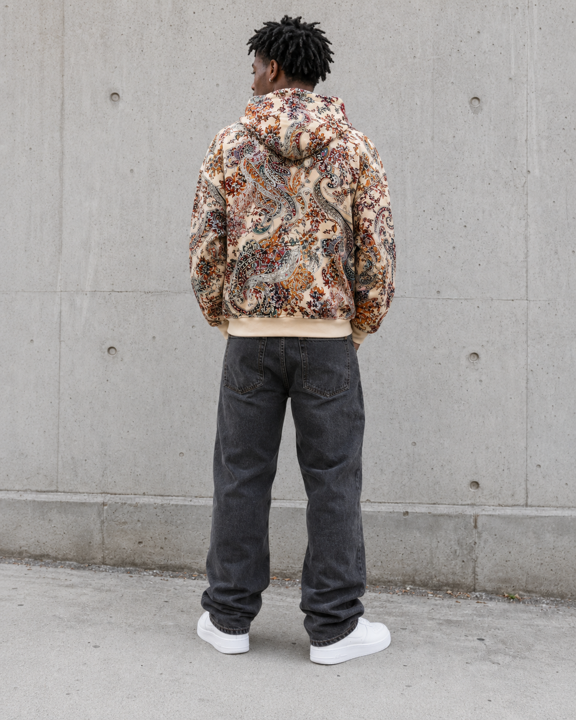 Person wearing a patterned hoodie and dark pants standing against a concrete wall.