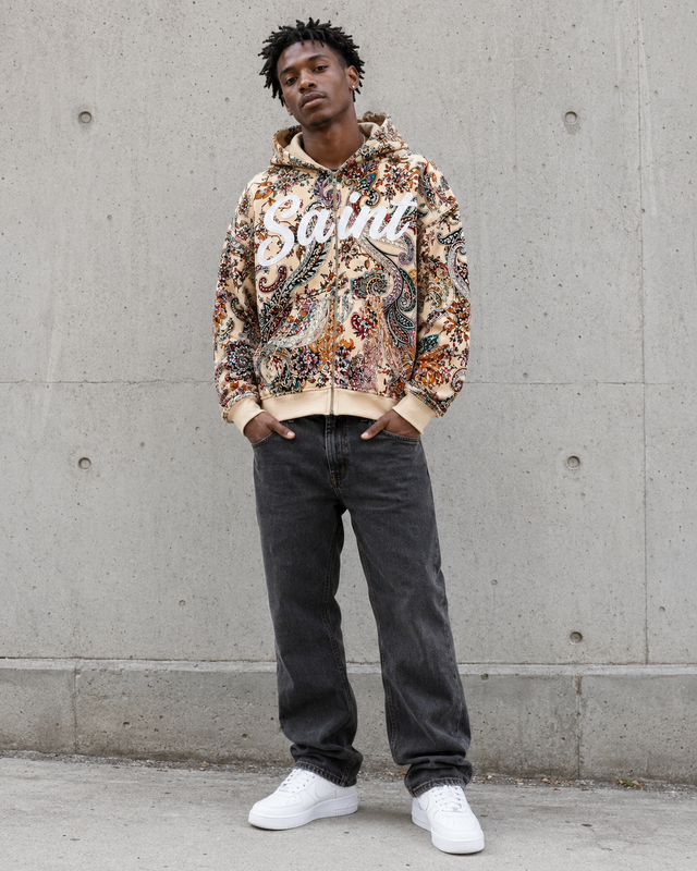 Person wearing a floral hoodie with text, dark jeans, and white sneakers against a concrete wall.