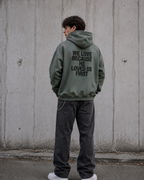 Person wearing a green hoodie with text on the back, standing against a concrete wall.