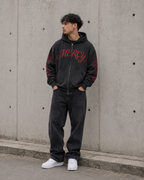 Person wearing a black hoodie with red text, dark pants, and white shoes against a concrete wall.