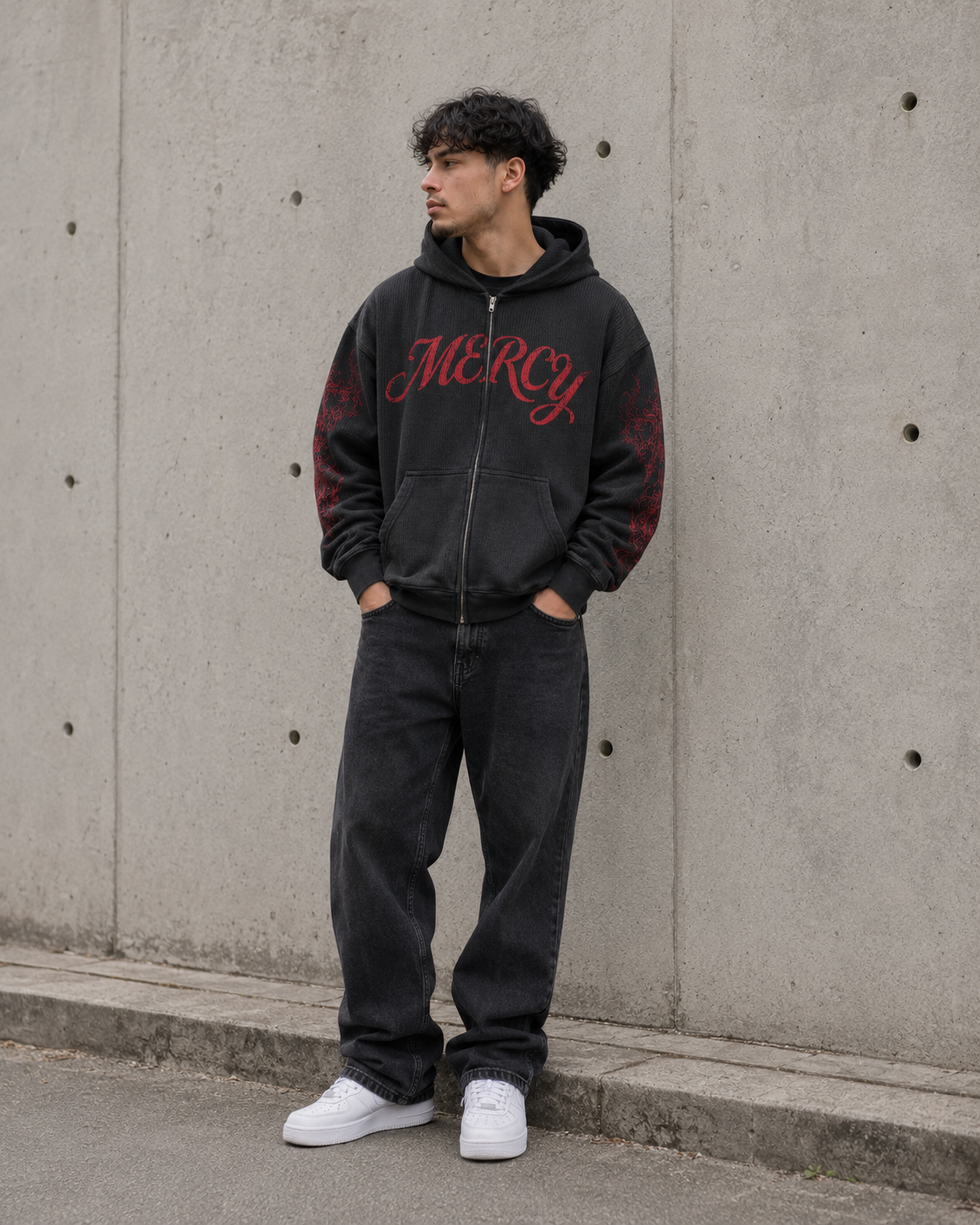 Person wearing a black hoodie with red text, dark pants, and white shoes against a concrete wall.