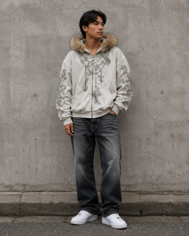 Person wearing a light-colored hoodie with fur hood and dark jeans standing against a gray wall.