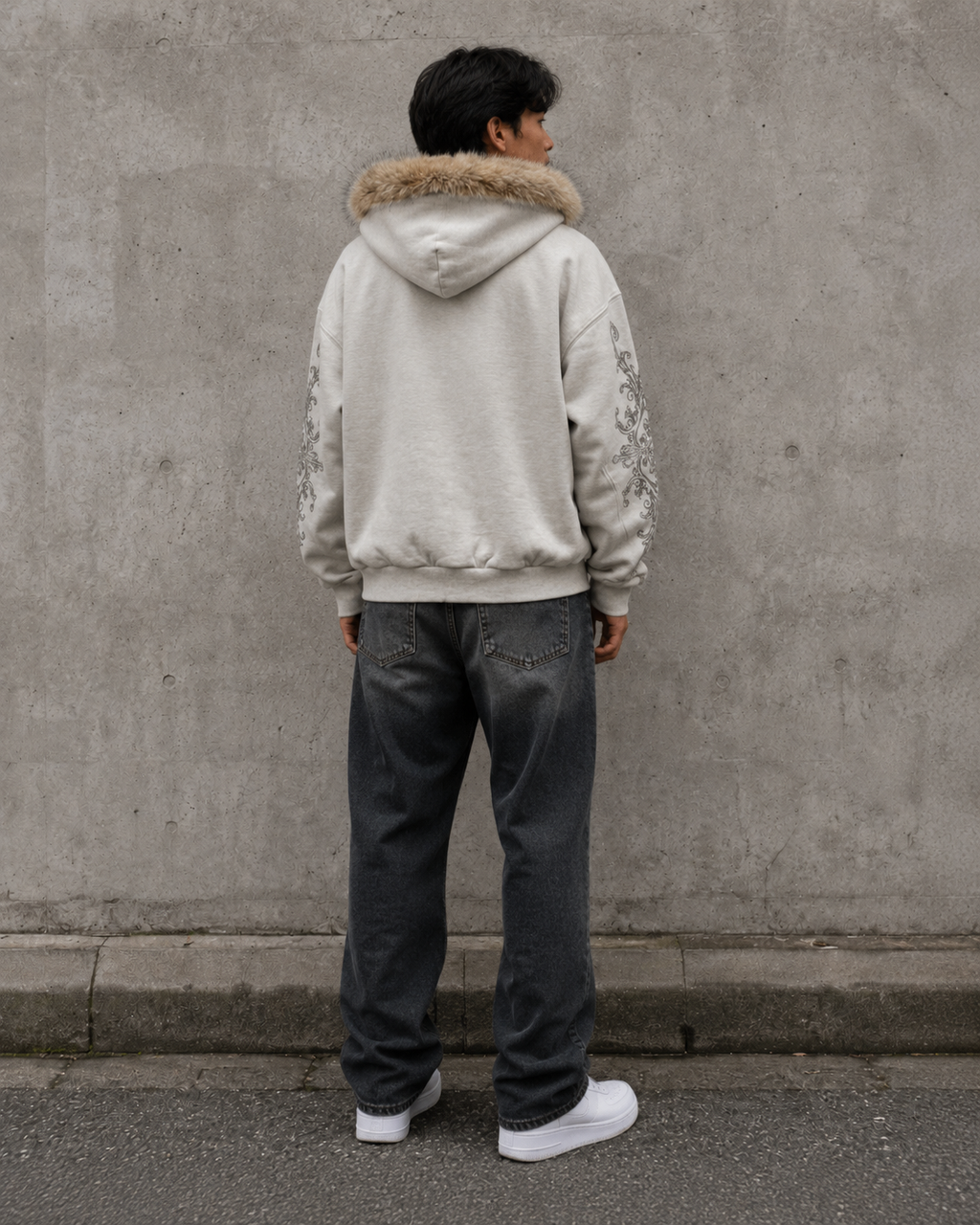 Person wearing a light-colored hoodie with fur hood and dark pants standing against a gray wall.
