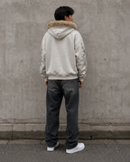 Person wearing a light-colored hoodie with fur hood and dark pants standing against a gray wall.