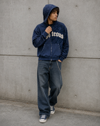 Person wearing a navy blue hoodie with text and jeans against a gray wall.