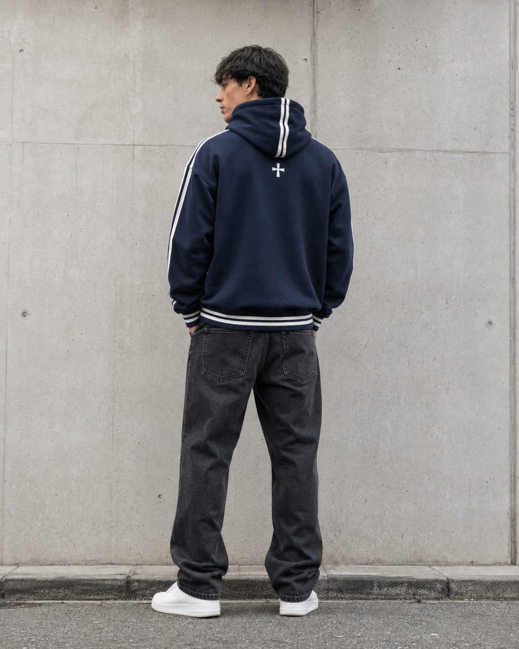 Person wearing a navy hoodie with white stripes and dark pants against a gray wall.