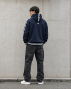 Person wearing a navy hoodie with white stripes and dark pants against a gray wall.