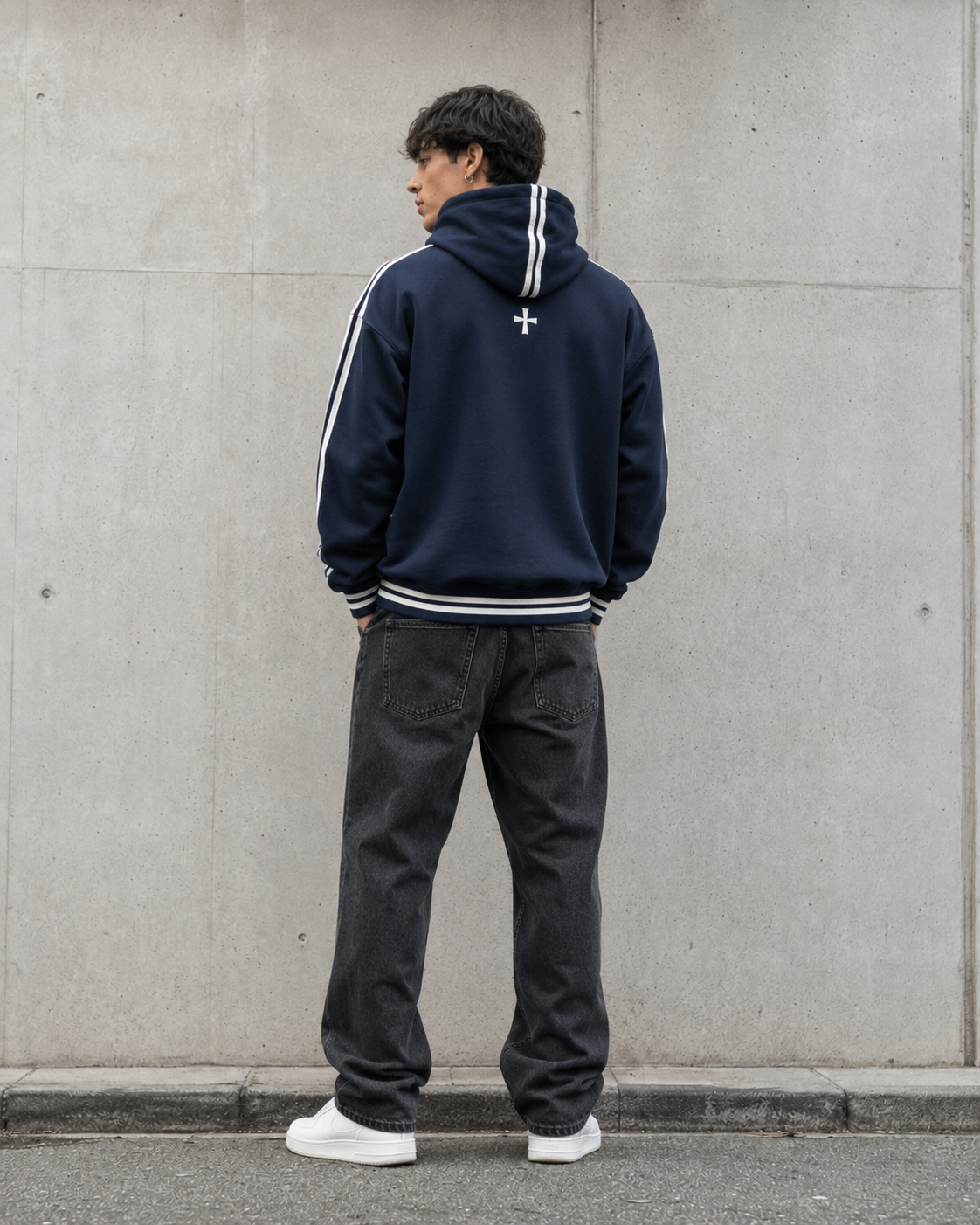 Person wearing a navy hoodie with white stripes and dark pants against a gray wall.