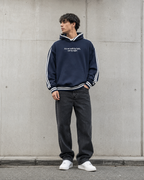 Person wearing a navy blue hoodie with white text and dark jeans standing against a gray wall.