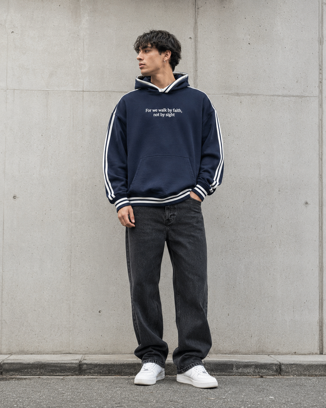 Person wearing a navy blue hoodie with white text and dark jeans standing against a gray wall.